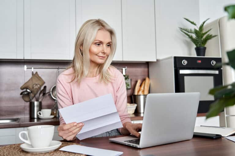 middle-aged woman holding paper looking at laptop